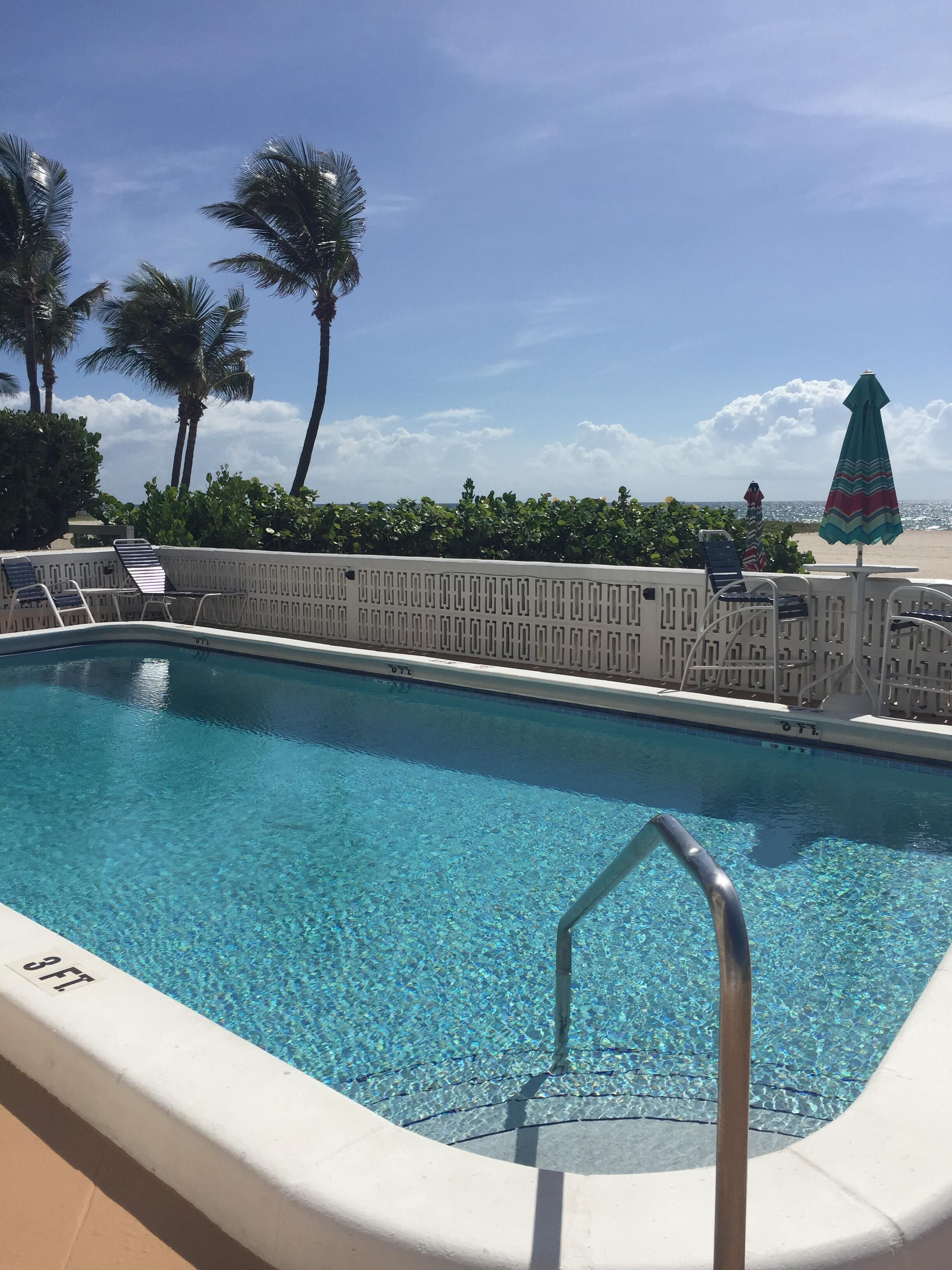 A swimming pool with clear blue water, surrounded by a white poolside area, palm trees, and a beach view.