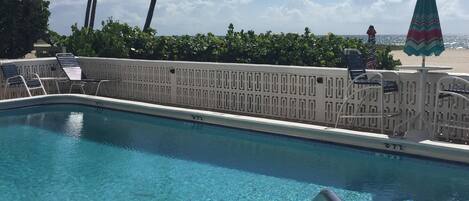 A swimming pool with clear blue water, surrounded by a white poolside area, palm trees, and a beach view.