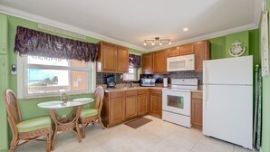 A compact kitchen with wooden cabinets, a white refrigerator, a microwave, a stove, and a dining area with wicker chairs. - DIRECTLY ON BEACH!  STUDIO Sleeps 2   Full Kitchen, Free parking, Heated Pool (Pompano Beach)