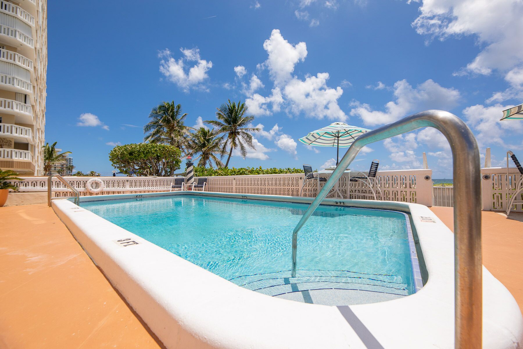 Une piscine avec un escalier en métal, entourée d’une terrasse blanche, de palmiers et d’un ciel bleu azur.