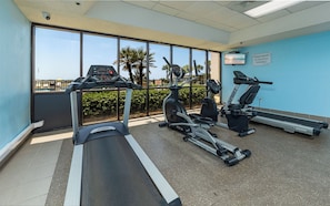 Ein Fitnessraum mit Laufbändern, Crosstrainern und Fahrrädern, mit Blick auf Palmen und einen Pool im Freien.