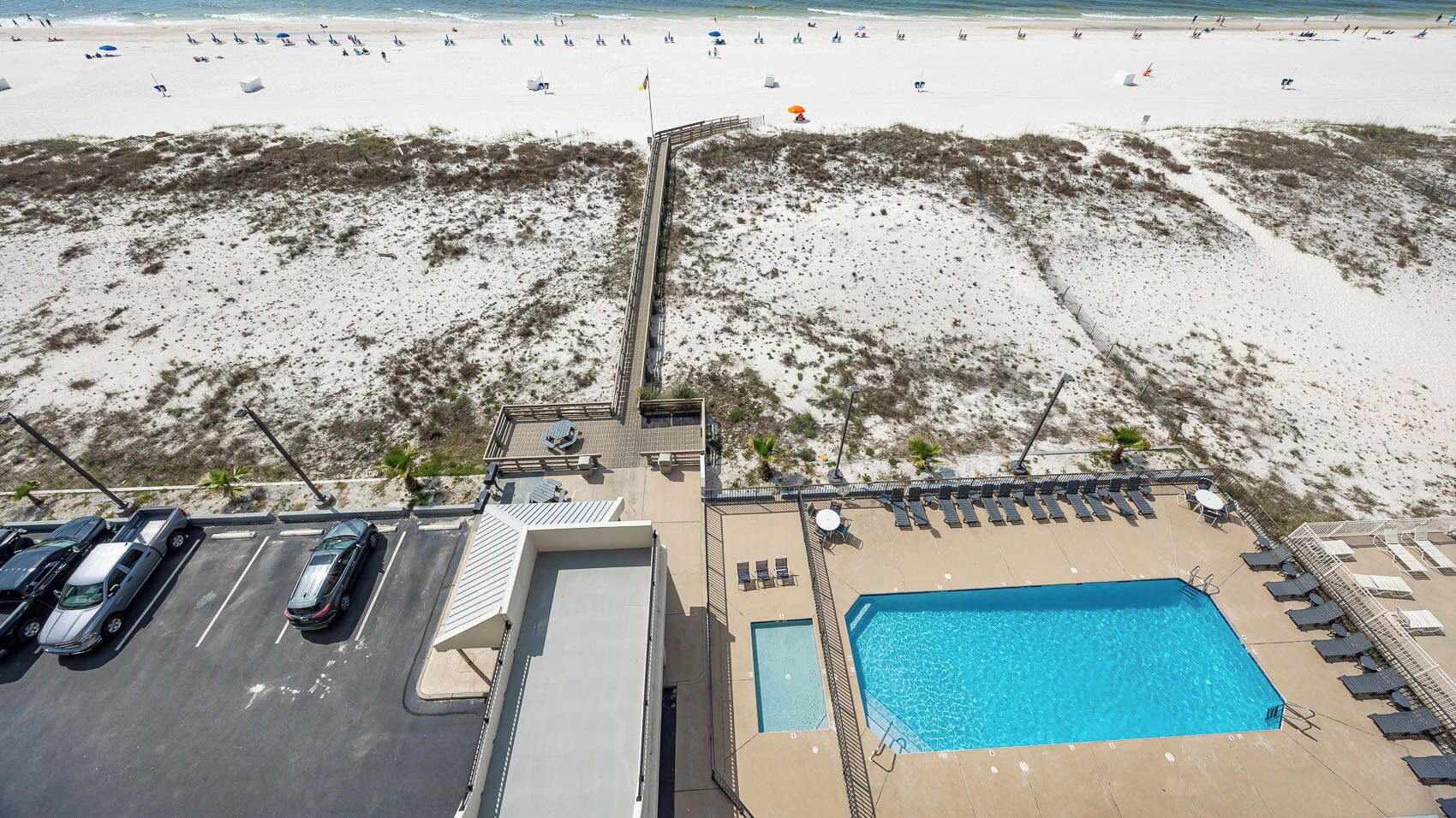 Vista aérea de un hotel frente a la playa con piscina, sillas de playa y un estacionamiento.