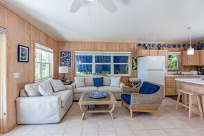 A cozy living room with a sofa, a wicker chair, and a coffee table. There is a kitchen area with wooden cabinets and a refrigerator.