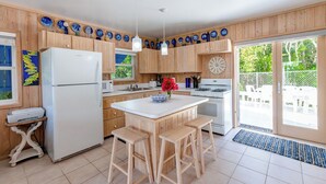 Una cocina con gabinetes de madera, un refrigerador blanco, una estufa y un lavamanos. Hay dos taburetes y un florero con flores en la encimera.