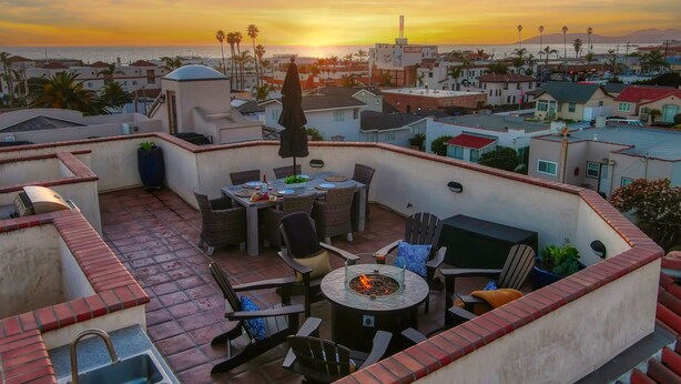 Une terrasse sur le toit, aménagée avec une table à manger, des chaises et un foyer, d’où l’on peut admirer le panorama urbain et l’océan au coucher du soleil.