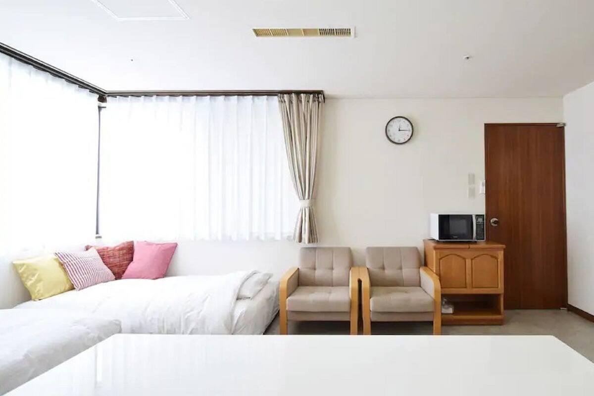 A hotel room with a bed, two chairs, a small wooden cabinet with a microwave, a clock on the wall, and a door.