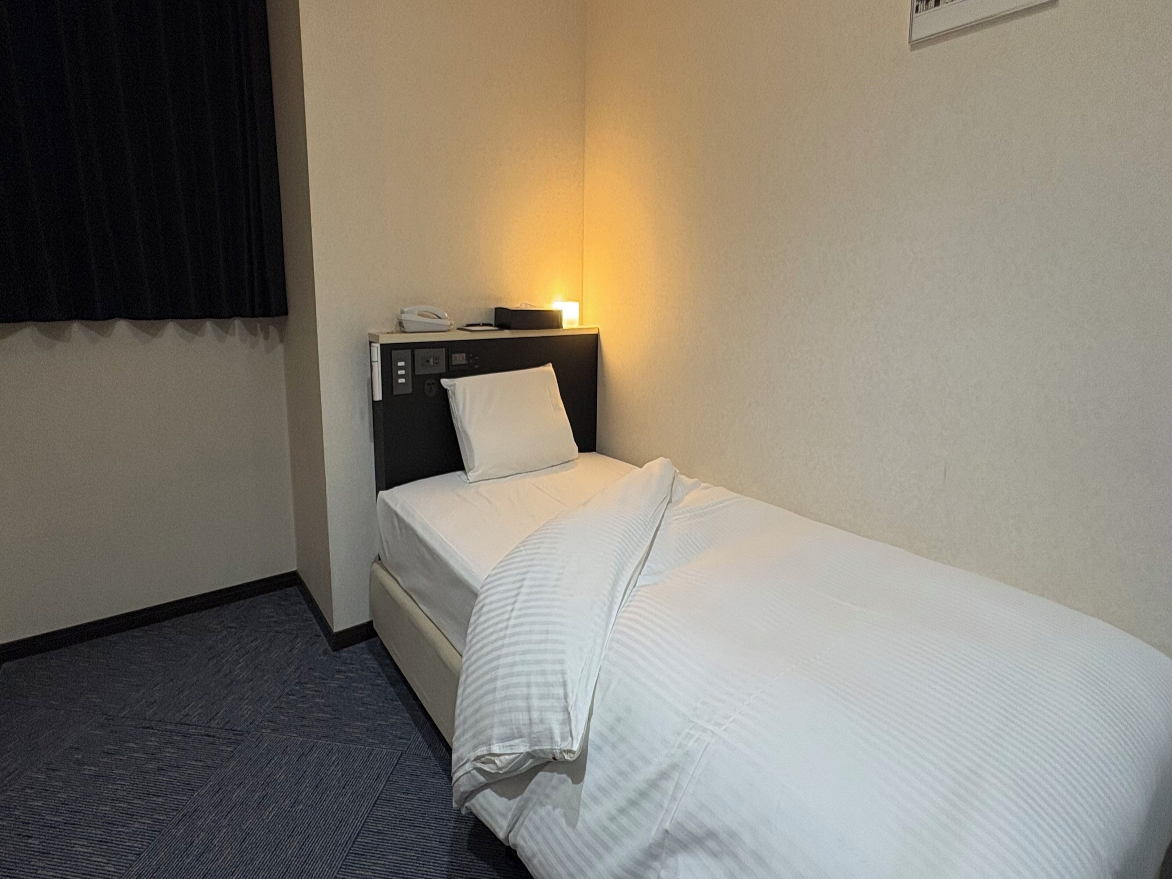 A hotel room with a single bed, a bedside table with a lamp, and a telephone.