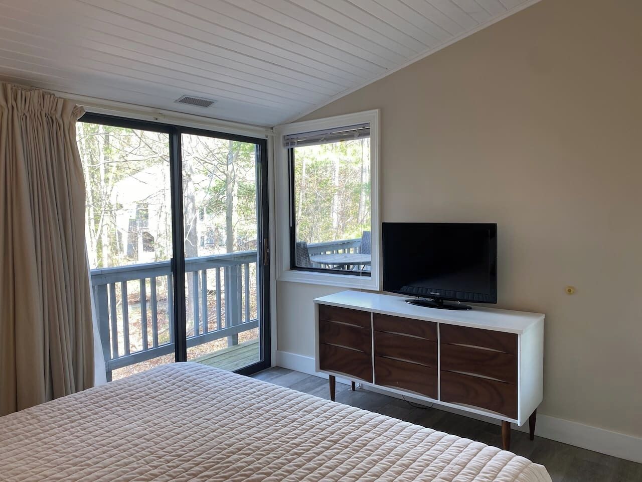 A bedroom with a large bed, a TV on a white cabinet, and a balcony with a view of trees and a deck.