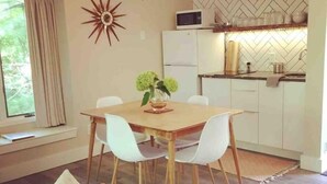 Un comedor con una mesa de madera y sillas blancas, un adorno en la pared y una cocina al fondo.