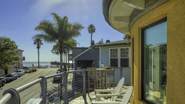 A balcony with chairs and a table overlooking a street with cars and palm trees. - Studio condo with ocean views, steps to the beach (Avila Beach)