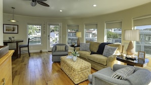 A spacious living room with hardwood flooring, a ceiling fan, a dining area with a table and chairs, and large windows offering views of the neighborhood. - Studio condo with ocean views, steps to the beach (Avila Beach)