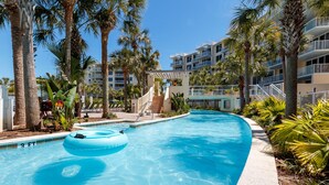 Une piscine avec un grand flotteur bleu, entourée de palmiers et bordée d’un immeuble à plusieurs étages doté de balcons.
