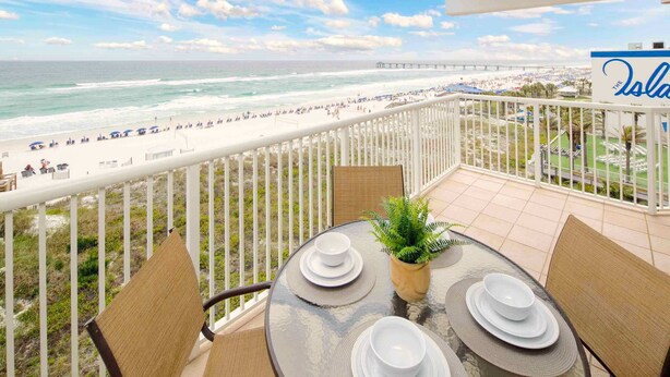 Un balcon avec une table en verre, dressée pour le dîner, offrant une vue sur une plage et une jetée.