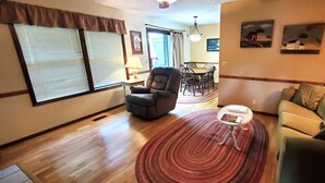 Una sala de estar con sofá, sillón, mesa de comedor y sillas. Hay una ventana con persianas, una lámpara y una obra de arte en la pared.