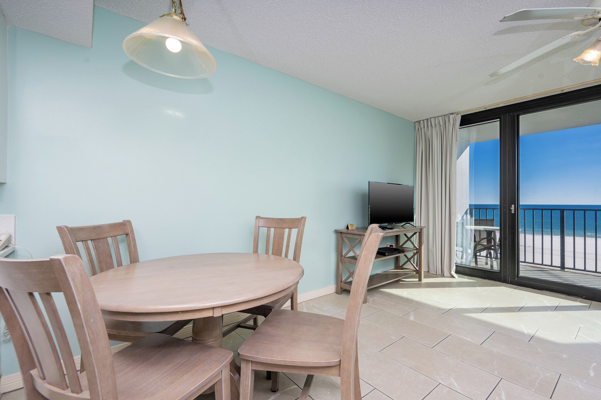 Un coin repas avec une table ronde et quatre chaises, un téléviseur posé sur un meuble, et un balcon avec vue sur la plage.