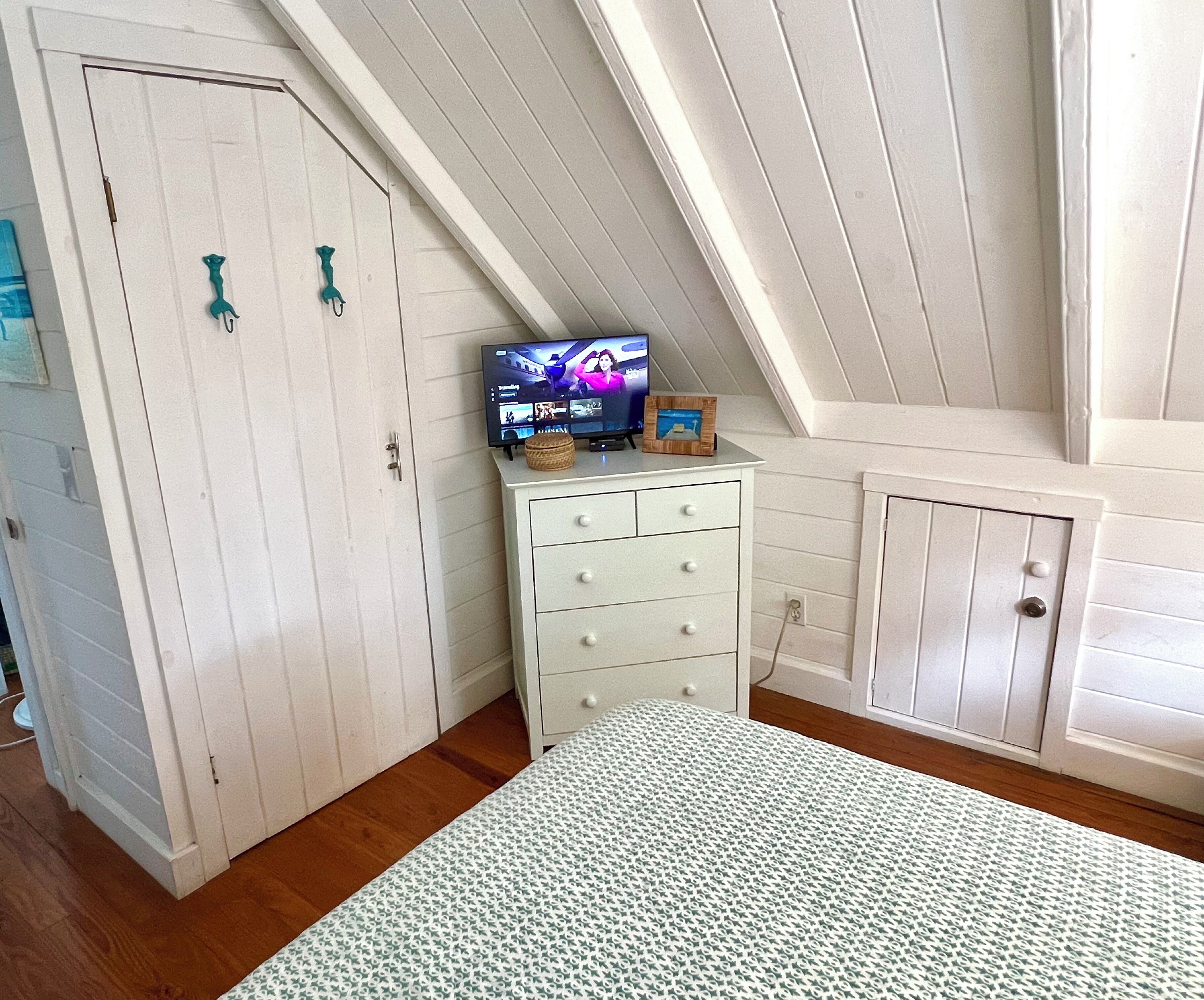 A bedroom with a sloped ceiling, a bed with a patterned bedspread, a dresser with drawers, a television on top, and a small decorative item on the dresser.