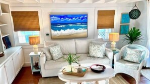 A living room with a sofa, coffee table, and decorative pillows. There is a painting of the ocean on the wall and a plant on the coffee table. - Dunwanderin-Historic Cottage in the Heart of Hope Town w/dock (Elbow Cay)