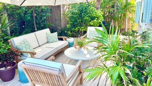 An outdoor patio area with a wooden deck, a white sofa with patterned cushions, a round table, and various potted plants. - Dunwanderin-Historic Cottage in the Heart of Hope Town w/dock (Elbow Cay)