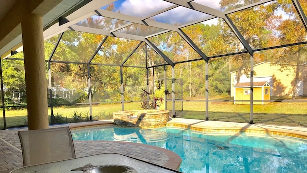 Une piscine extérieure couverte, dotée d’une table et d’une chaise en verre, entourée d’un jardin et offrant une vue sur une maison.