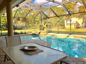 Piscina exterior cubierta con mesa y silla de vidrio, rodeada por un jardín y vistas a una casa.
