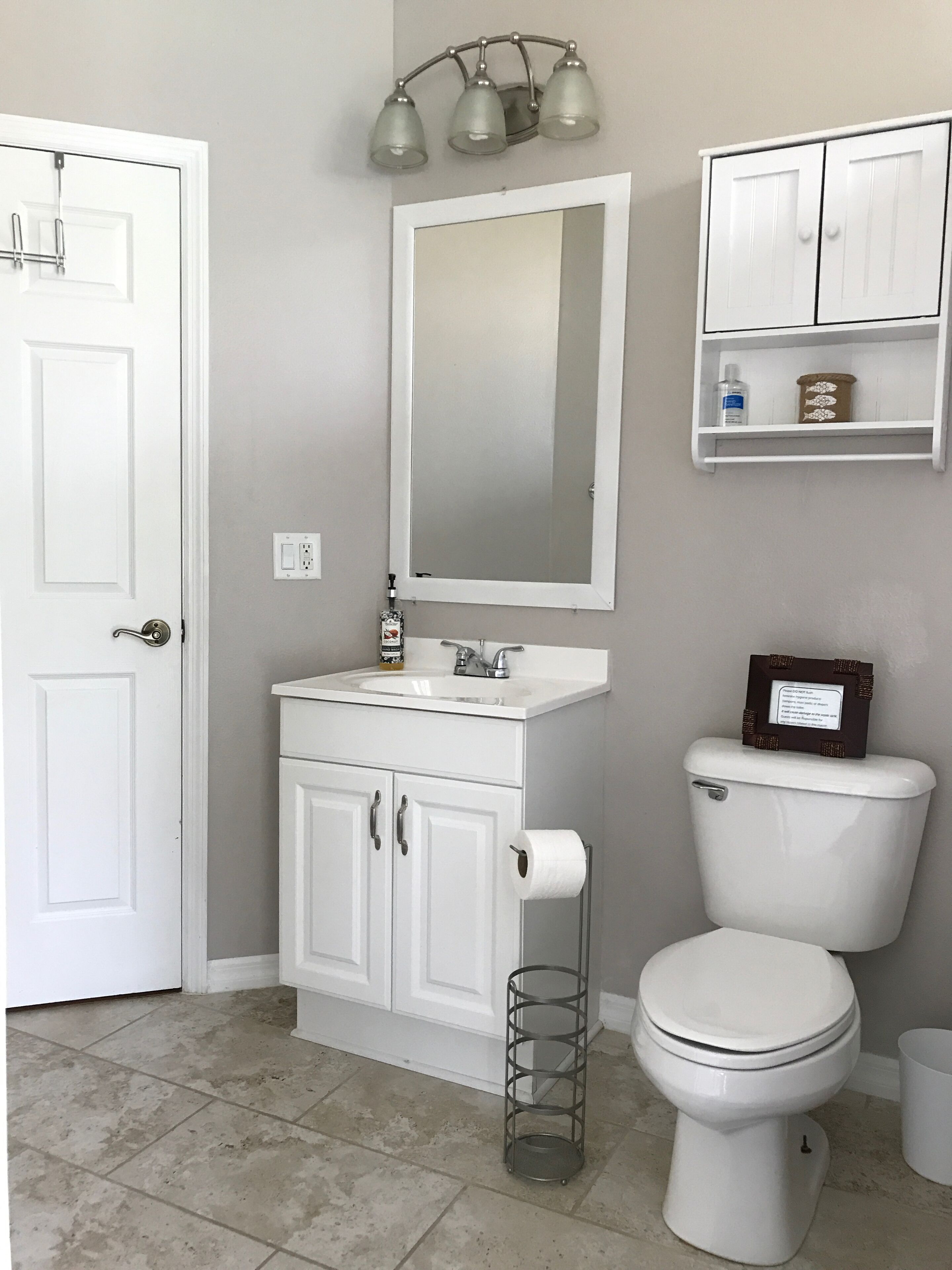 A bathroom with a white sink, mirror, and toilet. There is a shelf above the toilet and a door on the left.