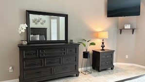 Une pièce avec une commode en bois foncé, une petite table d’appoint avec une lampe, une plante en pot et un téléviseur à écran plat fixé au mur.
