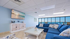 A modern living room with a large window, a blue sofa, a white coffee table, and a cabinet with decorative items. - *WEEKLY SPECIALS!* * Phoenix IV 2BR on 6th floor! (Orange Beach)