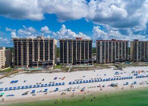 Un hôtel en bord de mer composé de plusieurs immeubles de grande hauteur, une plage de sable fin avec des parasols et des chaises longues, et un ciel bleu dégagé.
