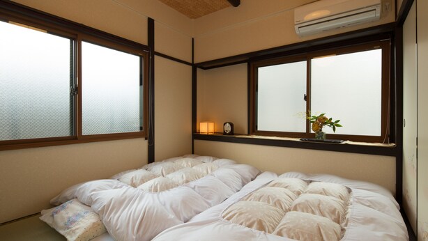 Una habitación con dos camas, un pequeño estante con un reloj y una planta, y ventanas amplias.
