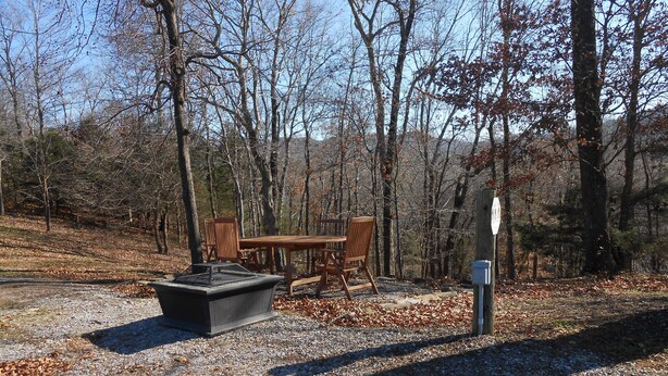 Un terrain de camping équipé d’une table et de chaises, d’un foyer et d’un poteau en bois portant une pancarte.