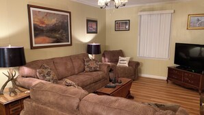 Un salon comprenant un canapé d’angle marron, deux fauteuils, une table basse, un téléviseur et un meuble en bois.