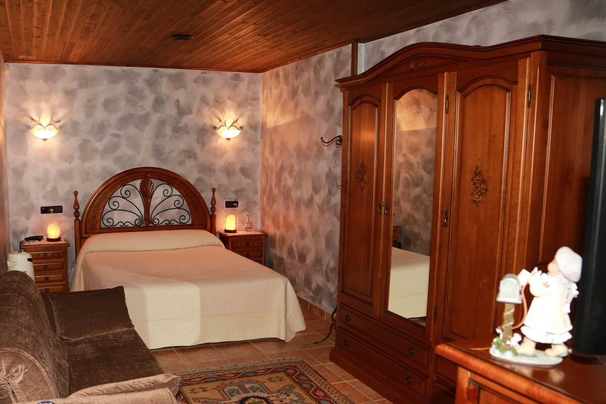 A bedroom with a wooden headboard bed, two bedside lamps, a wooden wardrobe, a nightstand, a brown sofa, a decorative globe, and a patterned rug.