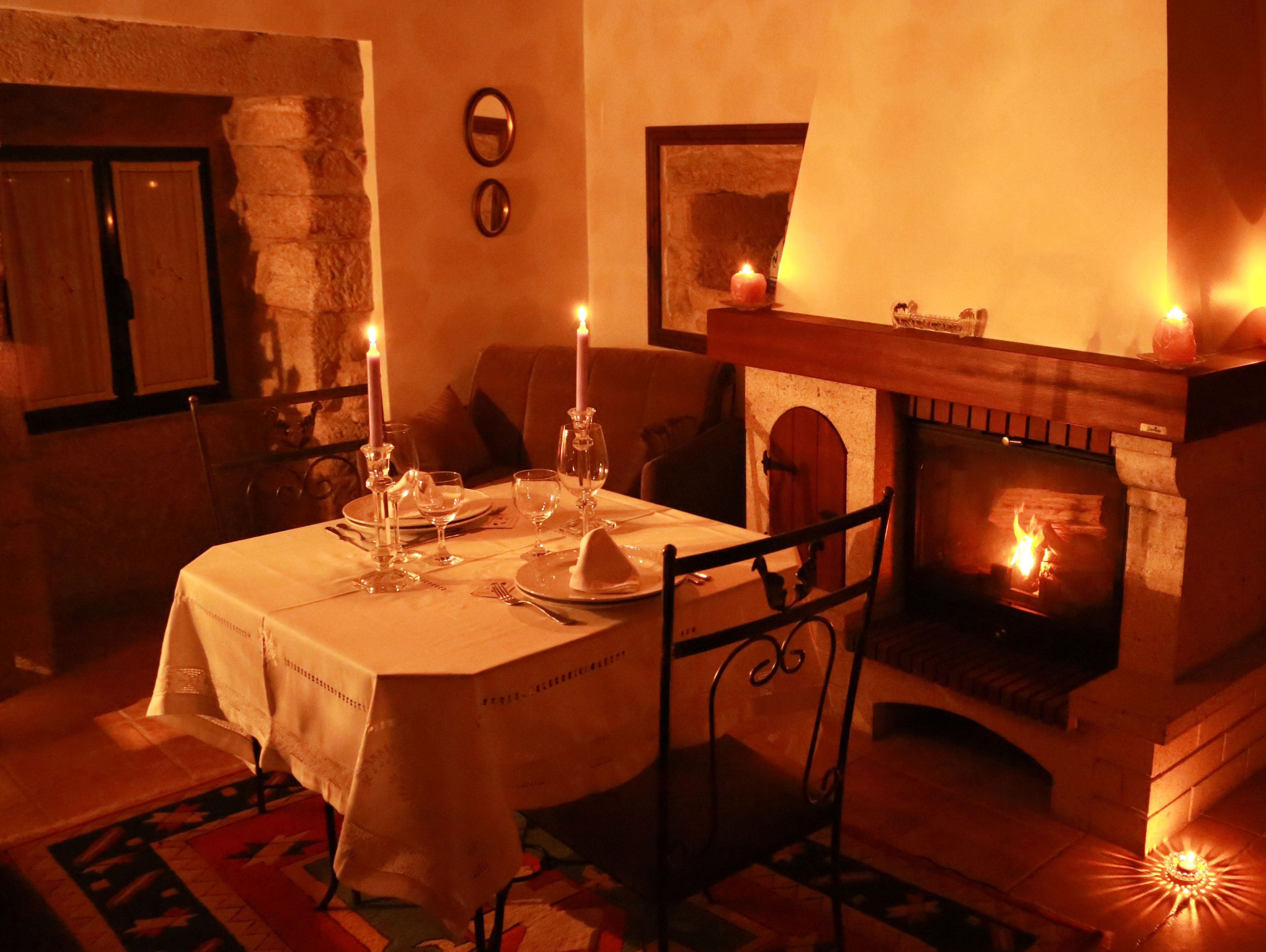Une salle à manger chaleureuse, éclairée d’une lumière douce, avec une cheminée, une table dressée pour deux personnes et un mur en pierre.