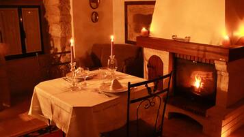 Une salle à manger chaleureuse, éclairée d’une lumière douce, avec une cheminée, une table dressée pour deux personnes et un mur en pierre.
