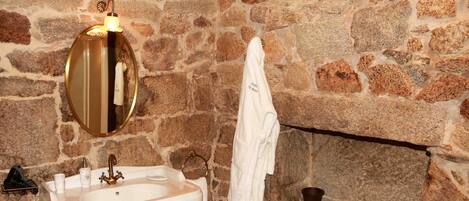 Un baño con pared de piedra, un lavabo blanco con grifo dorado, un espejo redondo y una toalla colgada en la pared.