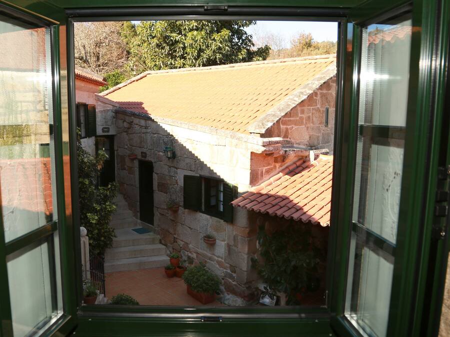 Vista pela janela de um edifício de pedra com telhado de telhas, uma pequena escadaria e plantas em vasos.