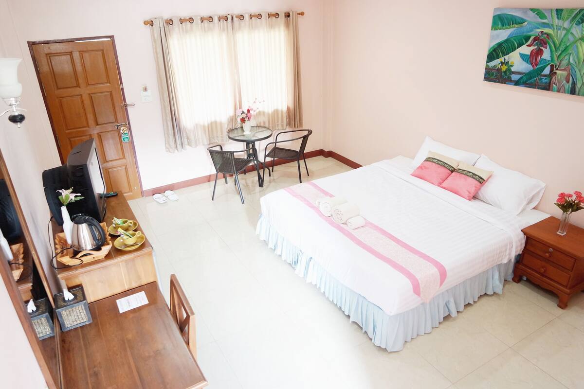 A hotel room with a bed, a desk with a chair, a small table with a vase, and a painting on the wall.