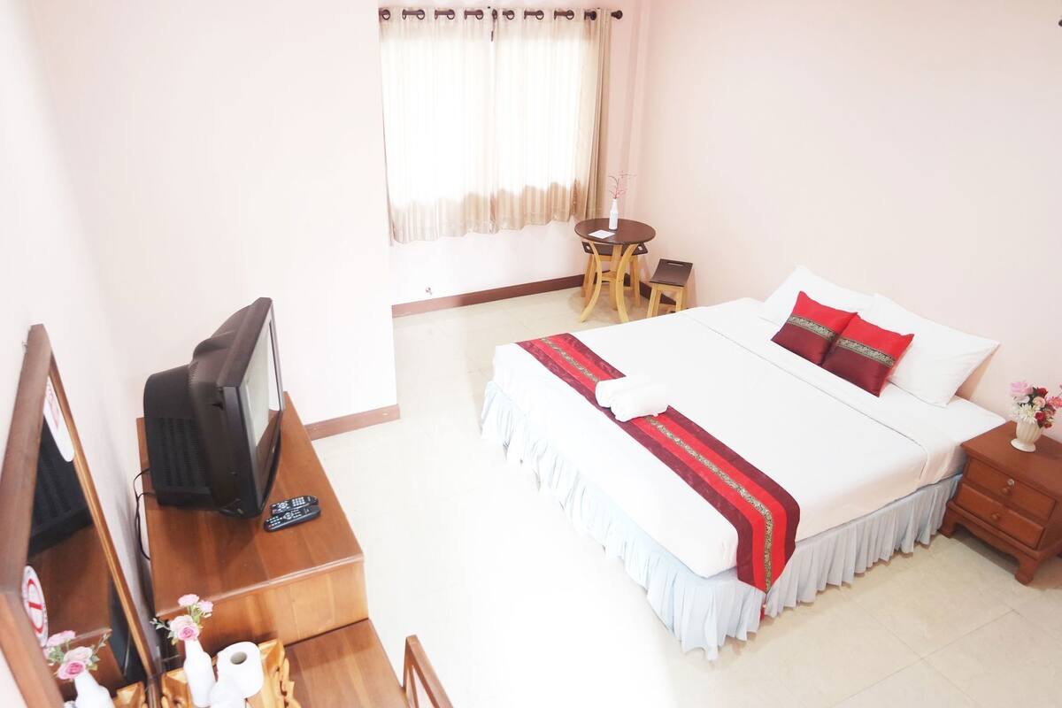 A hotel room with a bed, a desk with a television, a chair, a small table, and a window with curtains.