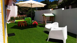 Un patio pequeño con césped artificial, una mesa con sillas, una casita de juegos y un paraguas blanco.