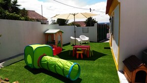 Un patio trasero cercado con césped artificial, una casita de juegos, una mesa con sillas y un pequeño refugio para perro.