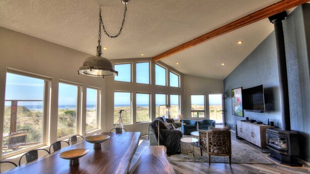 A spacious living area with a large wooden table, multiple chairs, a sofa, and a television mounted on the wall. - Oceanfront on Sandy Beach! Hot Tub! Dog Friendly! Direct Beach Access! Game Room! (Waldport)