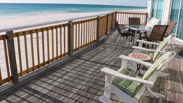 Um deck de madeira com vista para a praia, cadeiras brancas, uma mesa e guarda-corpo de vidro.