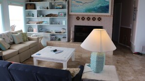 Uma sala de estar com uma mesa de centro branca, um sofá azul e um abajur verde-água sobre uma mesa branca. Há uma estante com vários objetos decorativos e um quadro sobre a lareira.