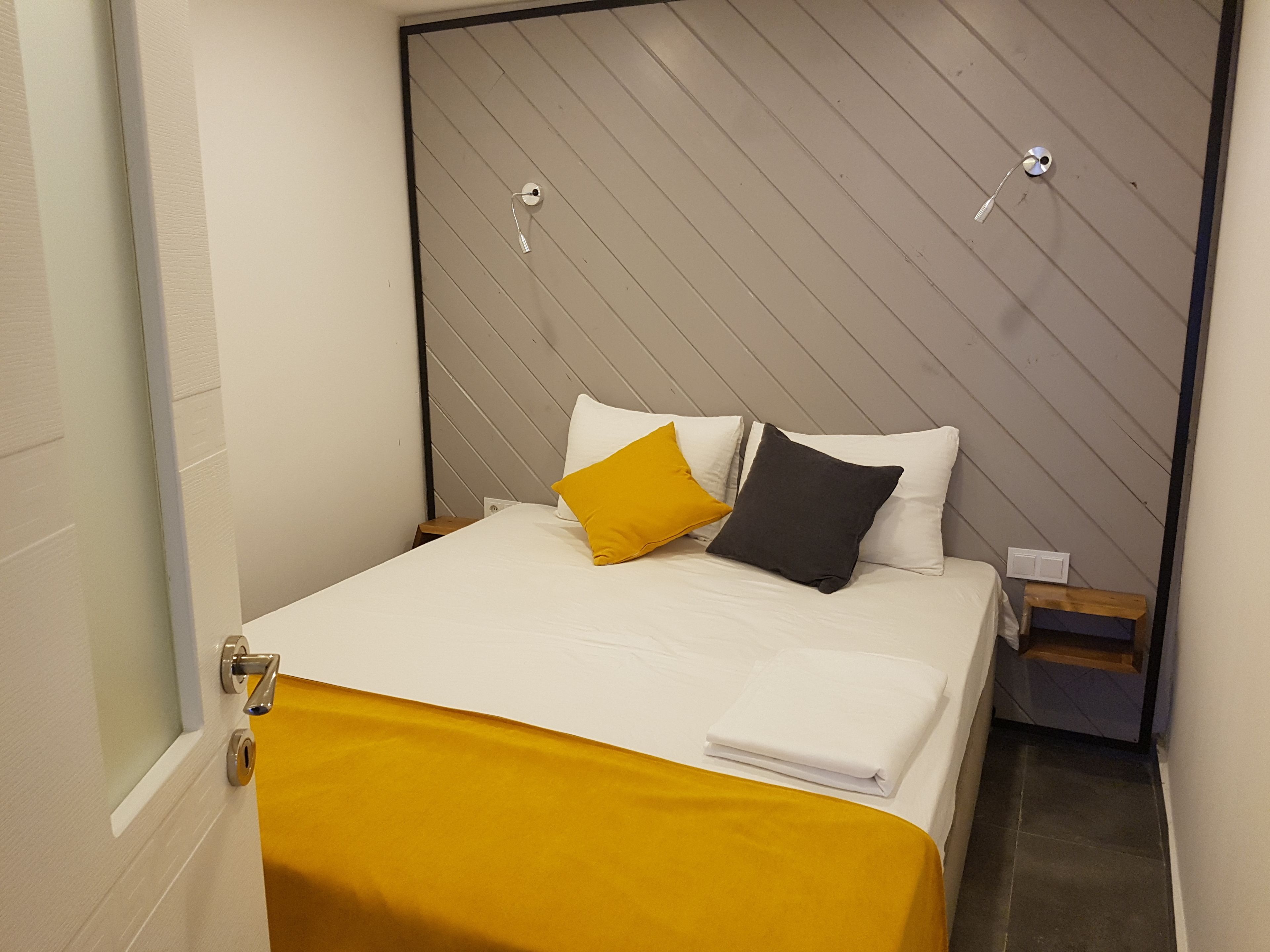 A neatly made bed with white bedding and two pillows, one yellow and one gray, in a room with a wooden slat headboard and a small wooden nightstand beside it.