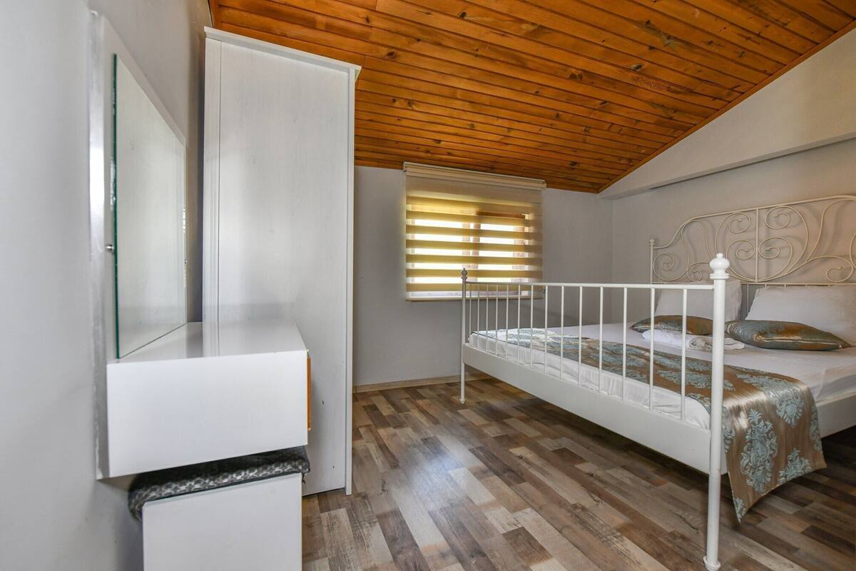 A bedroom with a wooden ceiling, a bed with a metal frame, a window with blinds, and a white wardrobe.