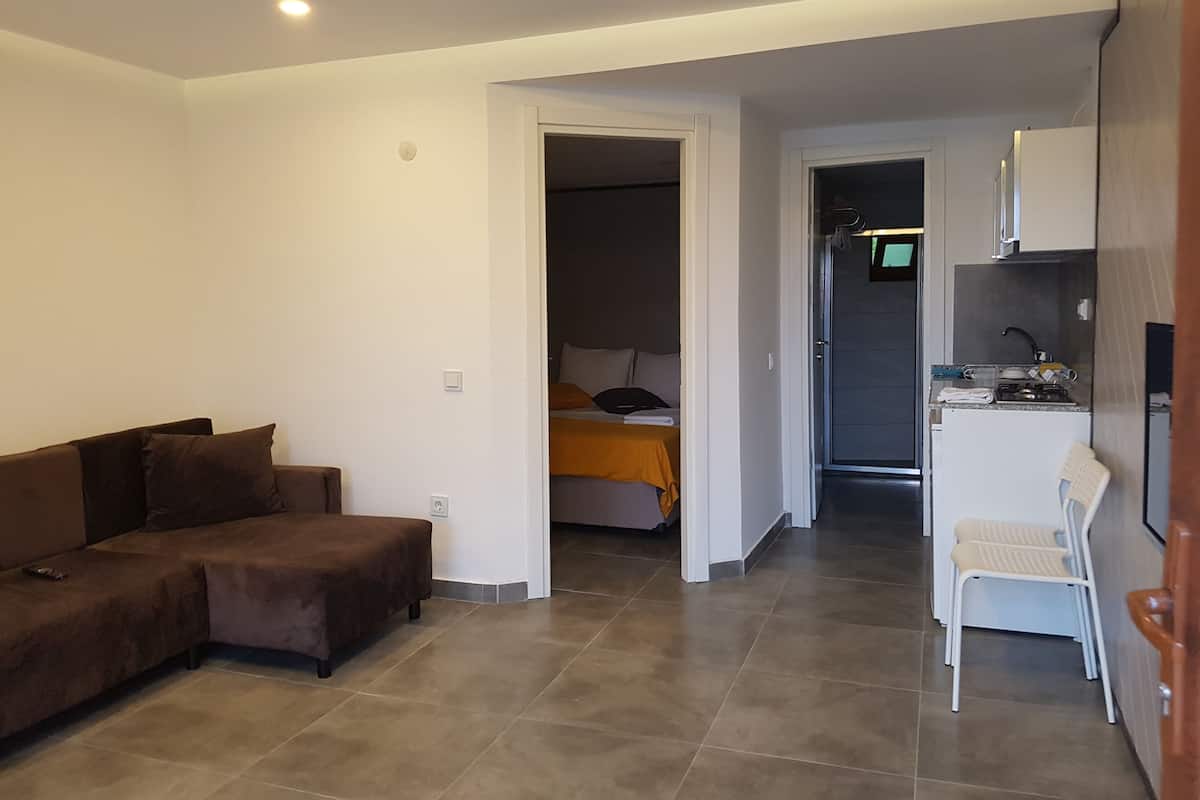 A compact living space with a sofa, a bed with an orange blanket, a kitchenette, and a small white chair.