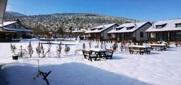 Un espacio exterior cubierto de nieve con bancos y mesas de madera, una pista de esquí y edificios con techos inclinados.