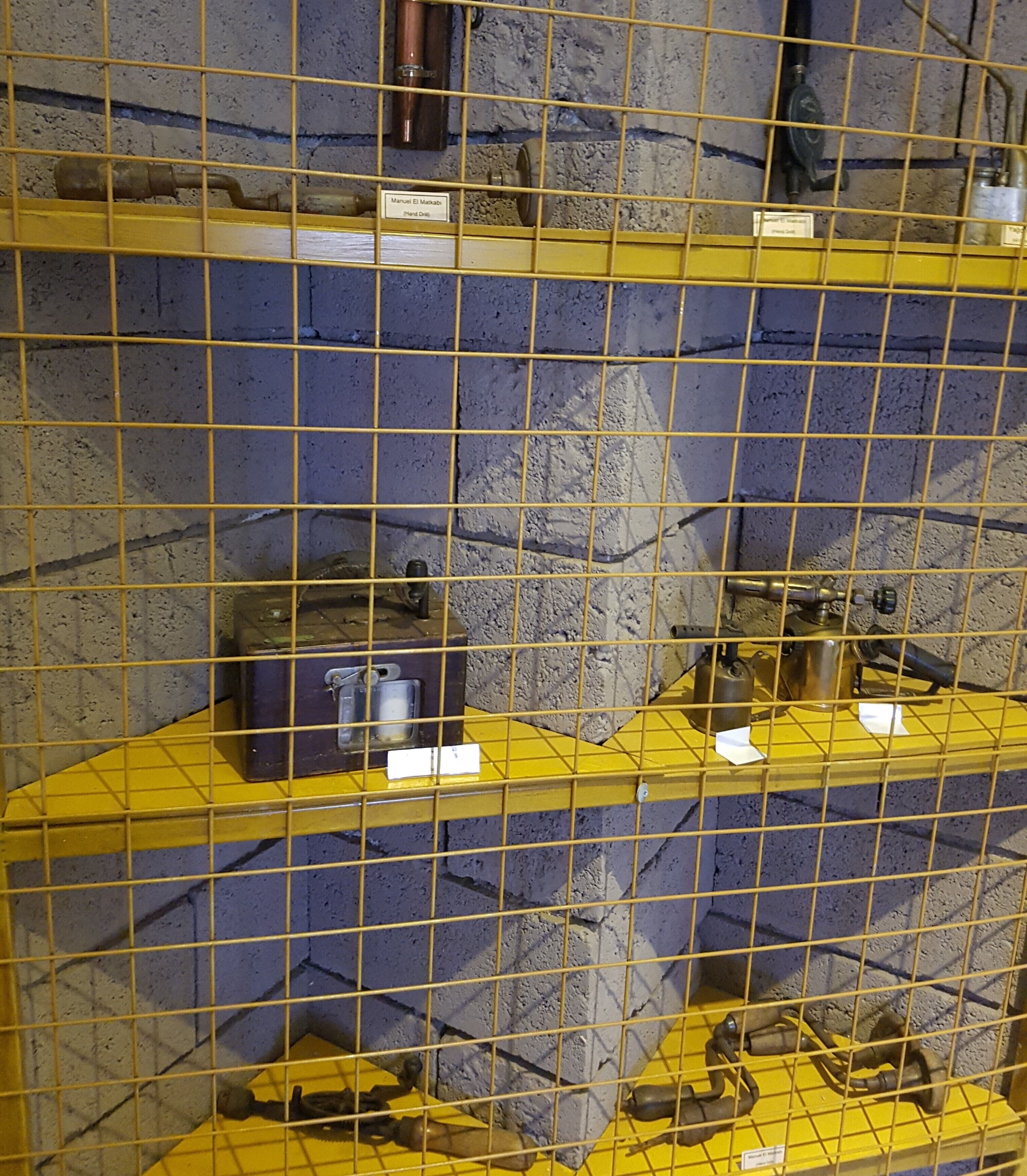 Un espace de stockage doté d’étagères jaunes et de grilles métalliques, contenant divers objets métalliques et des boîtes étiquetées.