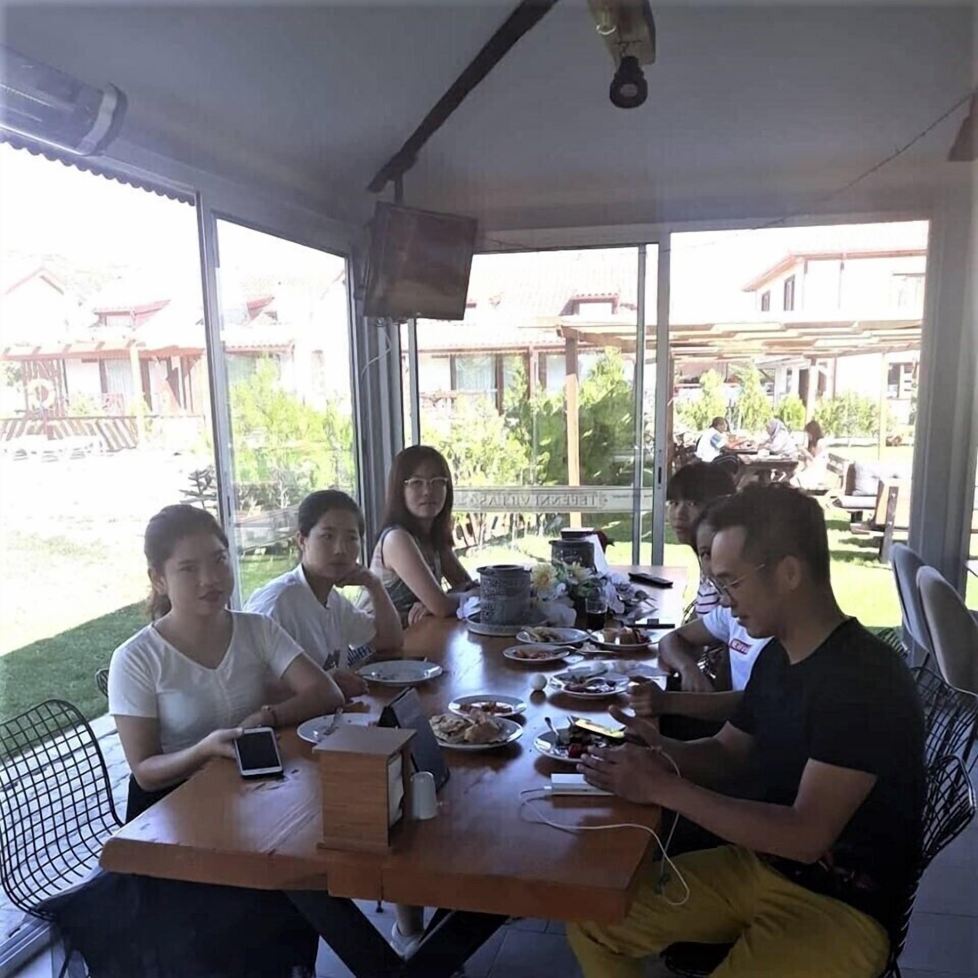 Un grupo de personas sentadas en una mesa de madera dentro de una habitación con ventanas amplias, una lámpara en el techo y vistas de un edificio.