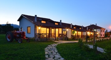 Una casa de dos pisos con un camino de piedra que lleva a la entrada, un tractor rojo estacionado al frente y un porche bien iluminado.